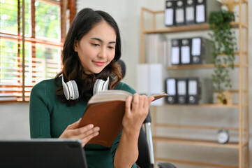 Fototapeta premium Relaxed young Asian female reading a book or her favorites novel in her office workplace.