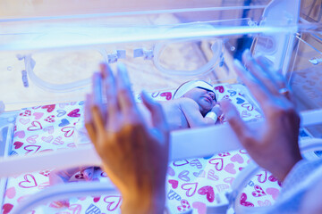 New born baby under the ultraviolet lamp in hospital. Unrecognizable mother hands on foreground. Selective focus.