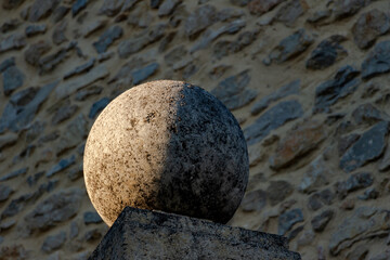 Stone sphere half enlighted by the sun, half in the shadow abd resting on a pillar. Stone wall in the background. Lussan, Gard, France.