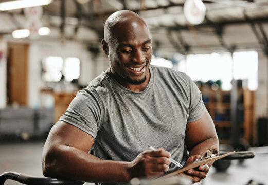 Gym, workout and personal trainer with checklist on clipboard consulting for training sports in gym. Black man of muscular, active and smiling fitness coach writing on health, wellness and exercise