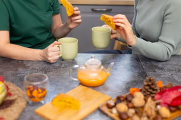 Two women in the kitchen are drinking sea buckthorn tea and eating pumpkin pie. People drink delicious healthy tea from green mugs. On the table stand a teapot with sea buckthorn tea, nuts and a pie