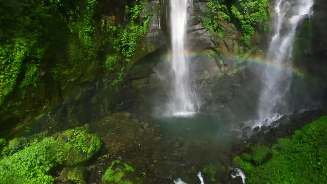 aerial of couple standing on a rock in the jungle at Sekumpul waterfall with a real rainbow in Bali Indonesia