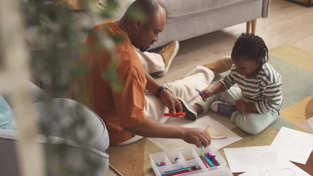From above shot of African American man and his pretty toddler daughter drawing rainbow with crayons sitting on colorful rug in living room