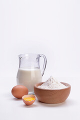 milk in carafe with broken egg and flour inside a wooden bowl on a white background