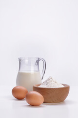 milk in carafe with eggs and flour inside a wooden bowl on a white background