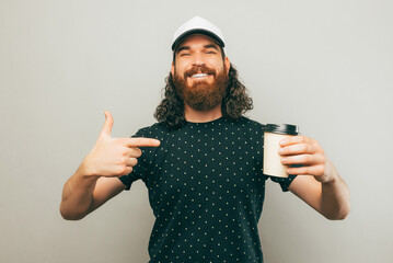 Hey look at this coffee, young bearded man pointing at paper cup of coffee to go over grey background. - obrazy, fototapety, plakaty