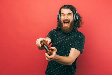 Photo of amazed bearded man playing games with joystick and screaming over pink background.
