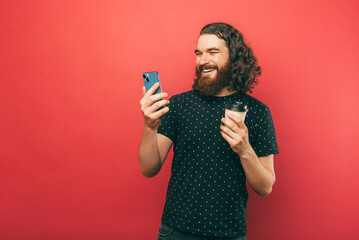 Photo of happy young handsome bearde man with long hair using smartphone and holding cup of coffee to go over pink background.
