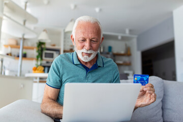 Fototapeta premium Mature man using laptop, holding plastic credit or debit card, senior grey haired customer making secure internet payment, shopping or browsing online banking service, entering information