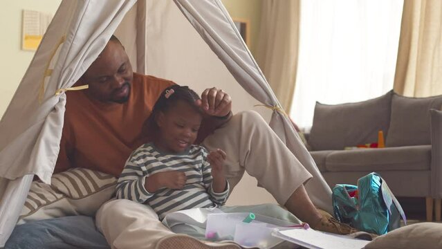 Happy Black man and his pretty 3 year old daughter spending leisure time together sitting in cozy handmade tent in bright spacious living room