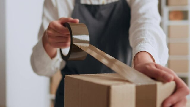 A warehouse worker in special clothes glues adhesive tape on a cardboard box while packing parcels for sending to customers of an online store of commerce, Girl warehouse worker of a transport company