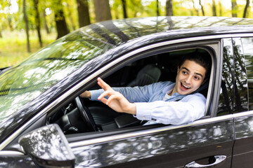 Portrait of young handsome brunette man driving car and greeting somebody with hand.