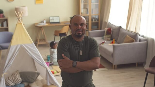 Medium slowmo portrait of African American solder in military uniform posing for camera with hands folded while staying at home with toddler daughter playing with toys in background