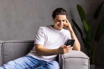 Fototapeta premium Portrait of an attractive smiling young man wearing casual clothes sitting on a couch at the living room, using mobile phone