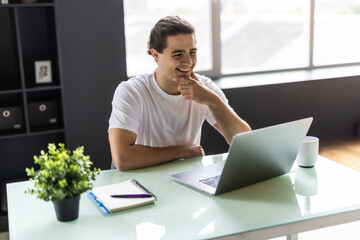 Smiling young man working on laptop in modern kitchen, checking email in morning, writing message in social network