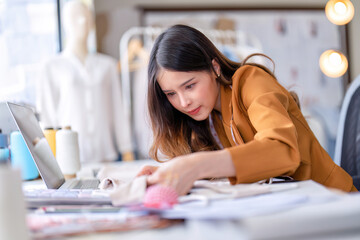 Young professional clothes fashion designer sitting near sewing machine use laptop computer and tablet pc to reference and concentrate on her work pattern