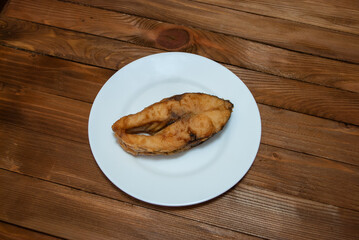 A piece of fried river fish on a white plate that stands on a natural wood table