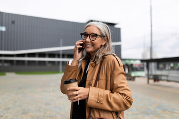 charismatic mature adult businesswoman with a cup of coffee and a mobile phone looks at the camera with a smile, business strategy concept