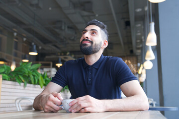 Fototapeta premium Portrait of happy positive Arab Muslim ethnic guy in glasses, young bearded man with black beard is drinking coffee sitting at table in cafe or restaurant, smiling
