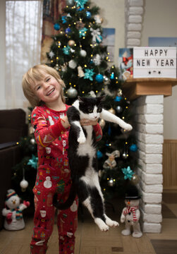 Cheerful Boy In Christmas Red Pajamas Holds A Domestic Black And White Cat In His Arms Stands At Home In Front Of The Christmas Tree. Concept Of Happy Cozy Family Winter Holidays Together With Pets