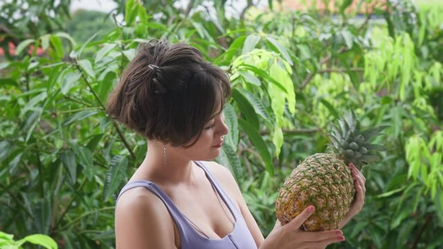 A happy Asian brunette girl with a short hairstyle holds a fresh pineapple, turns it over and rotates it. A woman touches pineapple leaves on a sunny day in a tropical garden.