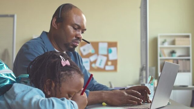 Serious African American businessman working on laptop in office while his pretty little daughter drawing him postcard sitting nearby