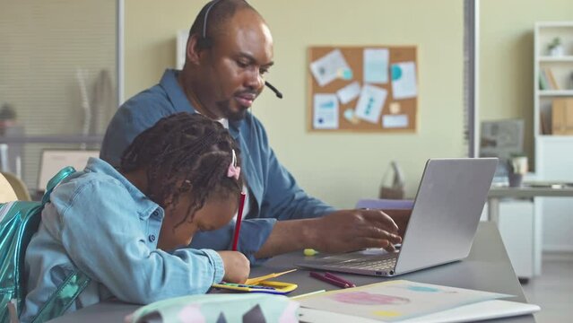 Cute 3 year old African American girl drawing while sitting at desk in her fathers office and her dad typing on laptop being busy with work