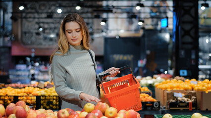 Young business woman girl buyer client blonde lady consumer stands in shop near counter with fruits in grocery store supermarket choosing juicy ripe apples for juice diet food buying puts in basket