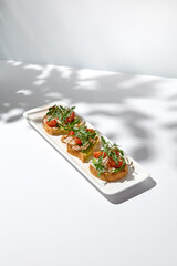 Aesthetic composition with shrimp bruschetta on white background with shadows from flowers. Italian bruschetta with shrimp, tomatoes and arugula on fine dining in summer. Elegant menu concept.