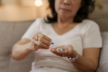 Elderly Southeast Asian woman holding heart disease pills sitting on the sofa alone, elderly health concept