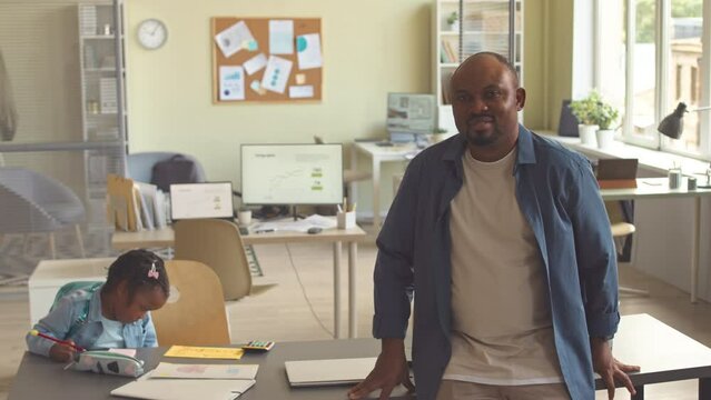 Medium slowmo portrait of Black man posing for camera while staying in office with his little daughter drawing at desk in background