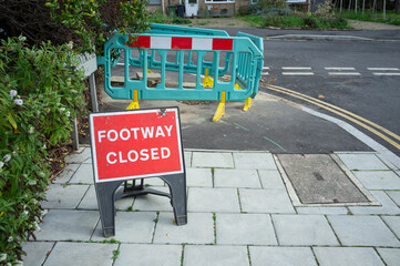 A red sign and barriers saying that footway ahead is closed as there are roadworks on the pavement....