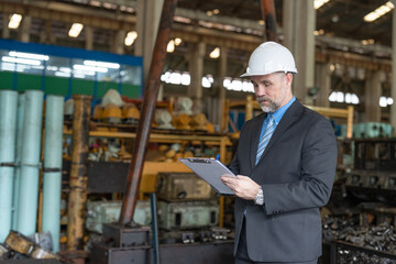 Senior businessman owner wearing helmet safety standing and writing on clipboard in heavy metal factory industry. Confident entrepreneur in suit checking lists on document at factory warehouse.