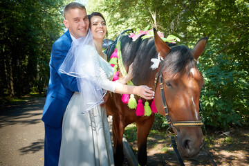 Stylish groom in a blue suit walk with the bride in a white dress in nature in the park with white horse. Wedding portrait of happy newlyweds on a background of greenery