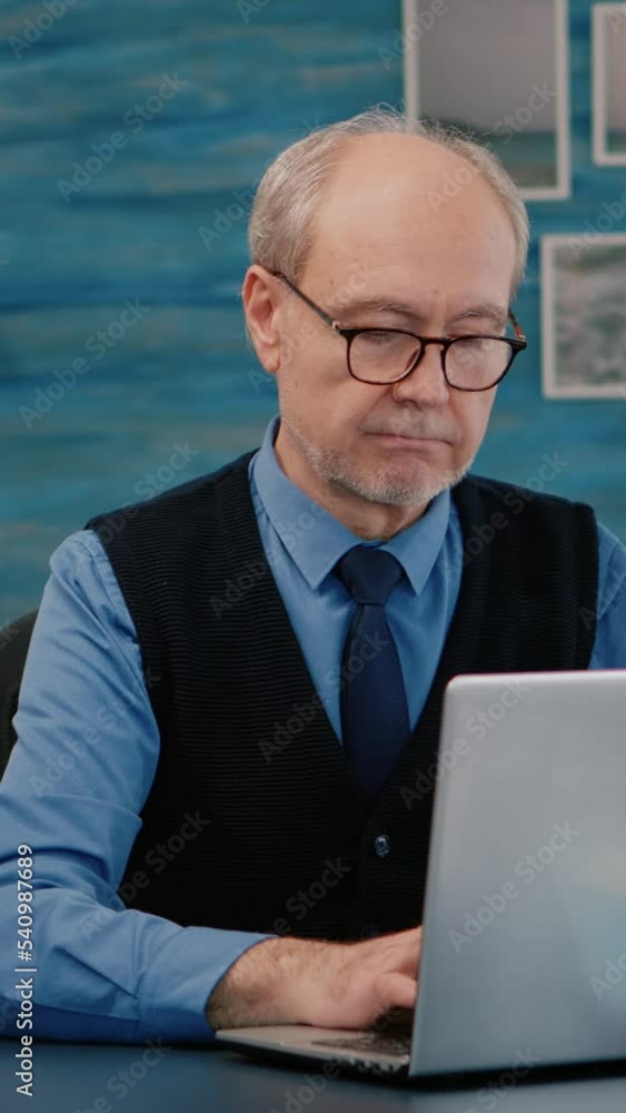 Poster vertical video: retired manager sitting at desk in front of camera smiling after typing at laptop wo