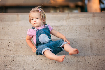 Young blonde in the rays of the setting sun. A cute little girl is sitting on the steps of the pier on the seashore. Street portrait of a little girl on a walk