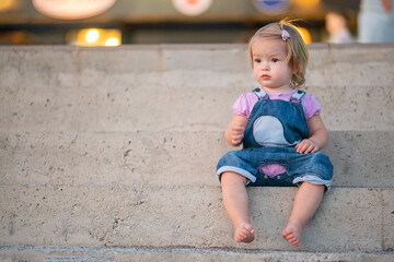 A cute little girl is sitting on the steps of the pier on the seashore. Young blonde in the rays of the setting sun. Street portrait of a little girl on a walk