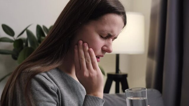 Young woman suffering from toothache, girl holding glass of water with ice touch cheek, face expression ache or feel pain, sensitive molar teeth, inflammation when drink cold, healthcare concept