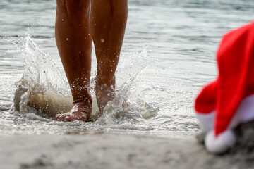 woman legs splashing on the shore with a Santa hat in the sand. christmas holidays