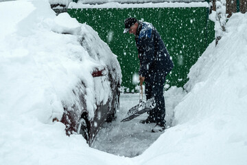 A man is digging up a snow-covered car with a shovel. Heavy snowfall. In the background a green...