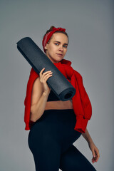 Pregnant woman holding yoga mat in hands, going for sport while awaiting baby, looking smiling at camera and touching her belly, standing against gray wall.