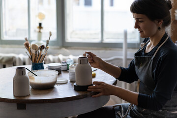 Young woman is engaged in favorite hobby drawing patterns on ceramic dishes sits at table in...