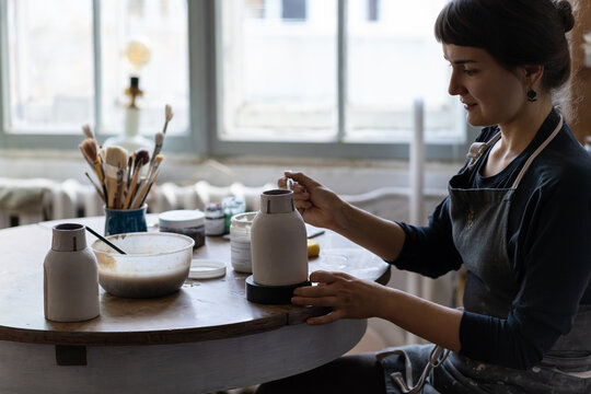 Young woman is engaged in favorite hobby drawing patterns on ceramic dishes sits at table in workshop. Self-sufficient girl artisan paints homemade vessels creating jugs for sale to collectors