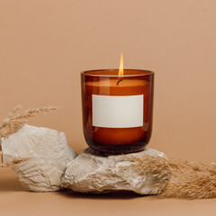 Burning handmade candle in a glass jar with empty label mockup, soy candle on stone podium with pampas grass, cozy interior, eco home decoration