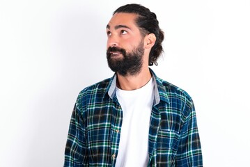 young bearded hispanic man wearing plaid shirt over white background with thoughtful expression, looks away keeps hands down bitting his lip thinks about something pleasant.