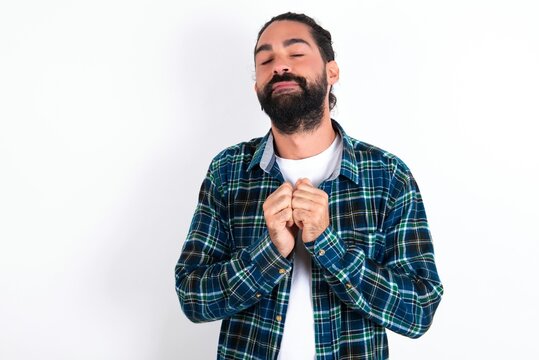 Dreamy charming young bearded hispanic man wearing plaid shirt over white background with pleasant expression, keeps hands crossed near face, excited about something pleasant.