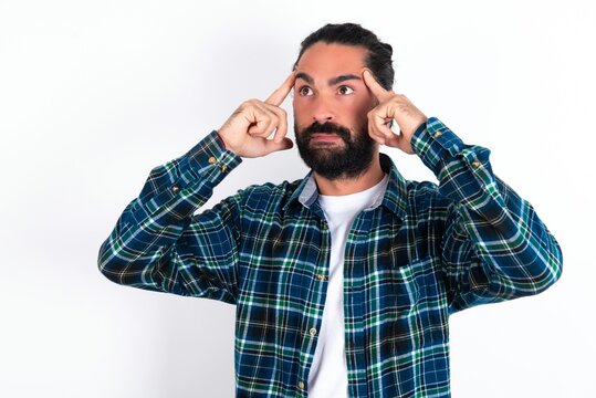 young bearded hispanic man wearing plaid shirt over white background with thoughtful expression, looks away, keeps hand near face, thinks about something pleasant.