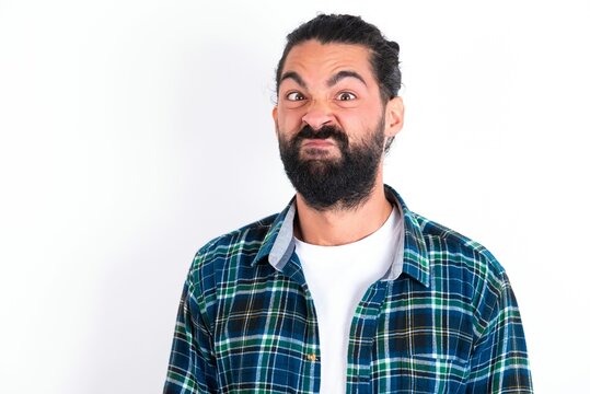 young bearded hispanic man wearing plaid shirt over white background crosses eyes, puts lips, makes grimace with awkward expression has fun alone, plays fool.