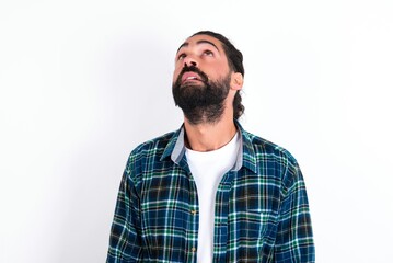 Fototapeta premium young bearded hispanic man wearing plaid shirt over white background looking up as he sees something strange.