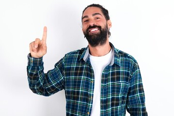 Fototapeta premium young bearded hispanic man wearing plaid shirt over white background showing and pointing up with fingers number one while smiling confident and happy.